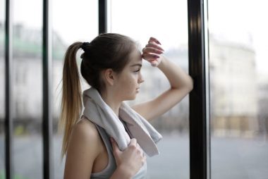 Side view of thoughtful young female athlete in sportswear with towel around neck standing near window with elbow on glass and looking away pensively while resting after training in modern gym