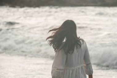 Woman in white dress walks by a stormy sea with waves crashing against the shore.
